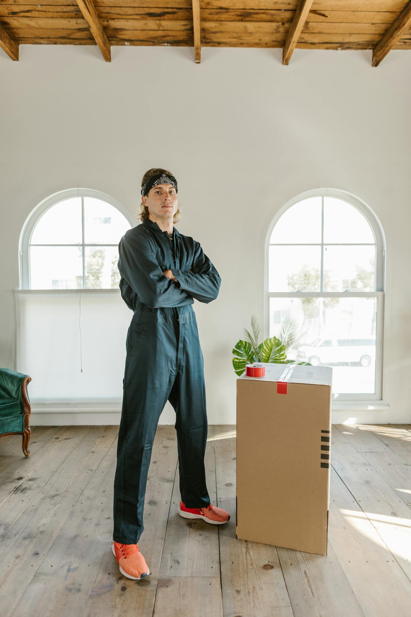 A young man in work attire stands confidently beside a moving box in a bright, wooden-floored apartment.
